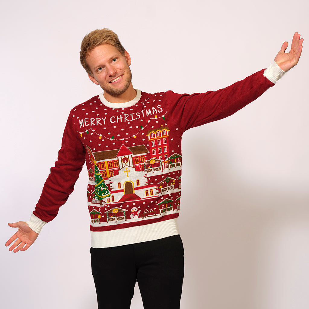 En poserande man klädd i en röd jultröja med en snötäckt stad på.