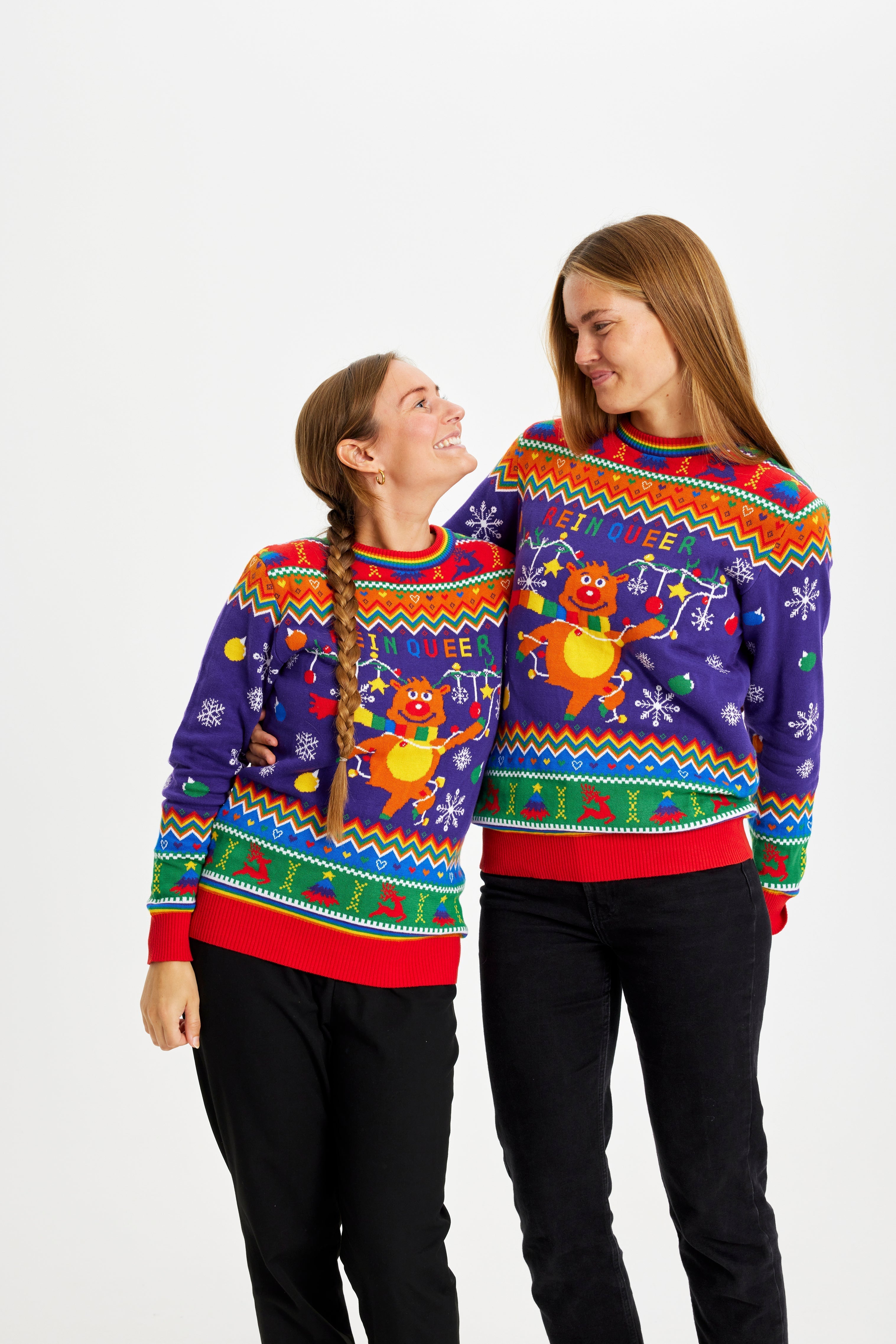 Två poserande kvinnor bär en jultröja med regnbågsfärger och citatet 
