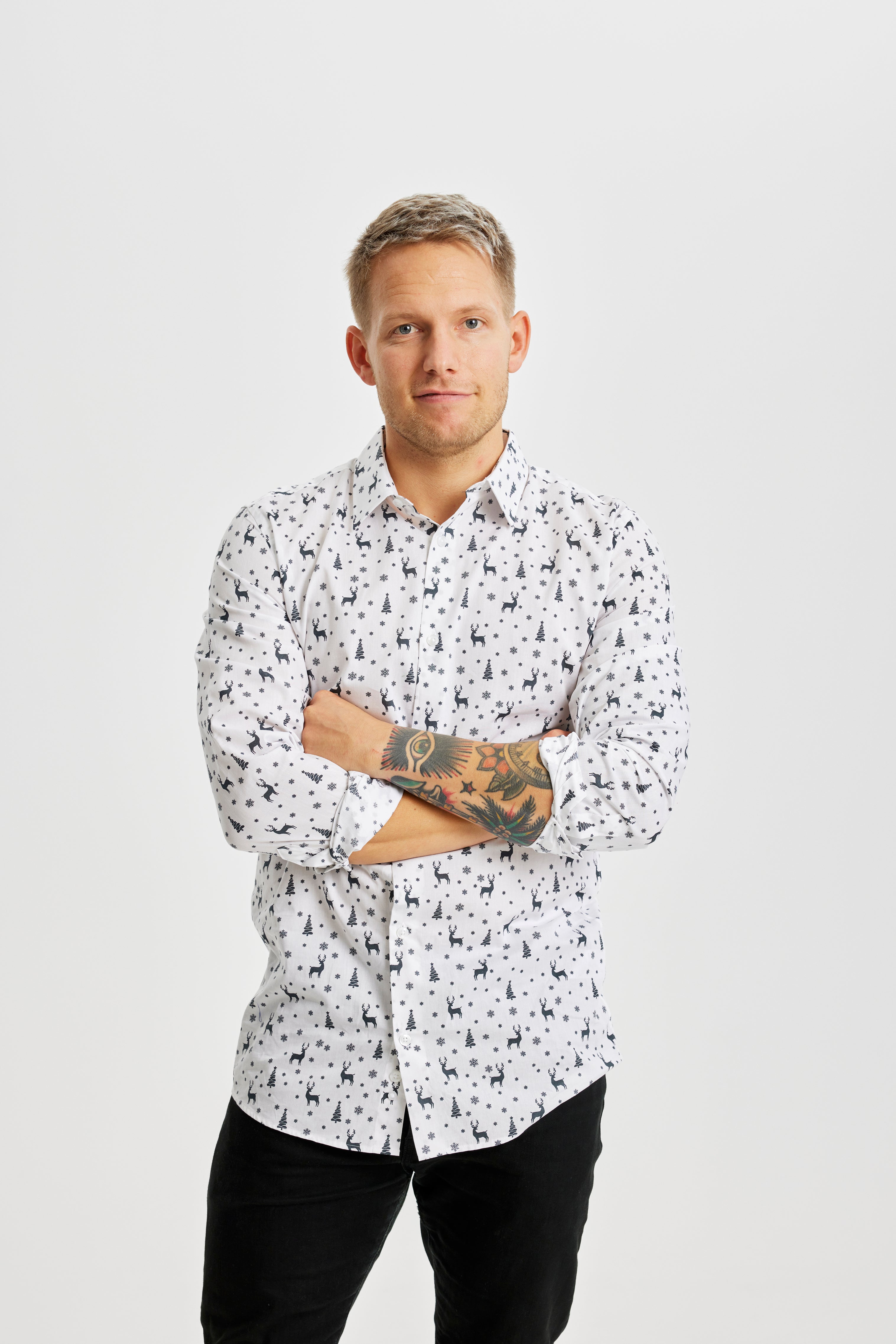 En leende man med armarna i kors klädd i en vit jultröja med julgranar och renar på.