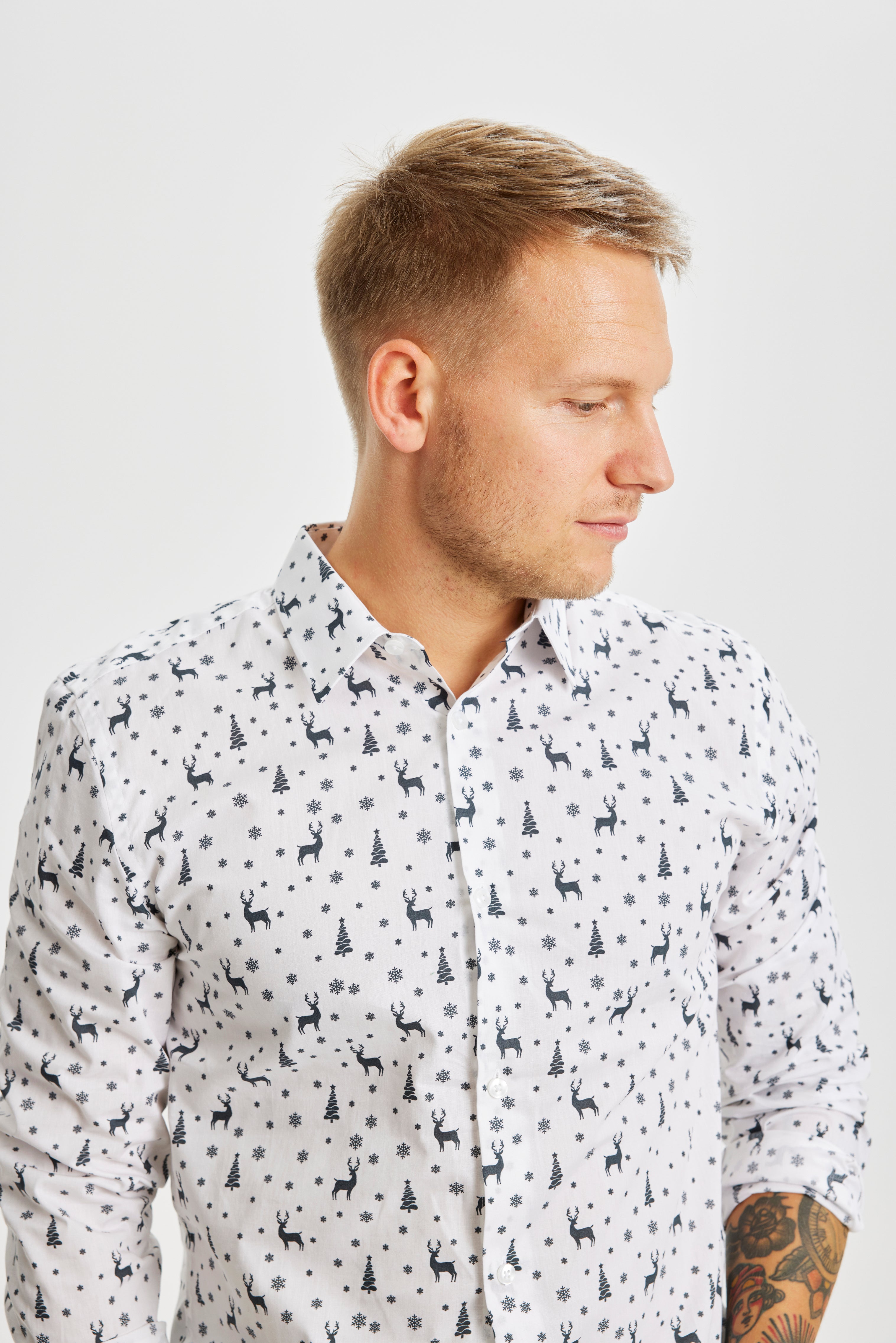 En man tittar åt sidan iklädd en vit julskjorta med julgranar och renar på.